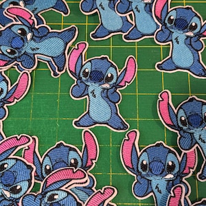 May include: A collection of blue and pink embroidered patches featuring the character Stitch from the animated film Lilo & Stitch. The patches are in various poses, with a white border, and are arranged on a green cutting mat.