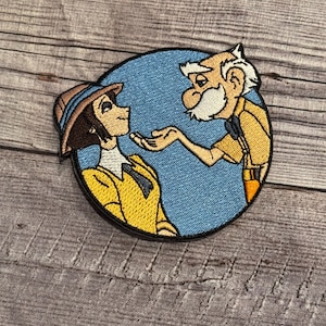 May include: Embroidered patch featuring a cartoon man and woman. The man is wearing a yellow shirt and brown pants. The woman is wearing a yellow jacket and a brown hat. The background is blue.