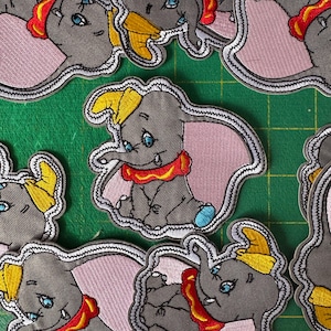 Könnte beinhalten: Gestickte Aufnäher mit der Figur Dumbo aus dem Disney-Zeichentrickfilm. Die Aufnäher zeigen Dumbo in Grau mit gelbem Hut, rosa Ohren und rotem Kragen. Die Aufnäher sind mit einem weiß-grauen, genähten Rand umrandet.