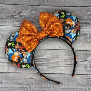 May include: A pair of orange sequined Minnie Mouse ears with a black headband. The ears are covered in a fabric print featuring characters from the cartoon show "Kim Possible".