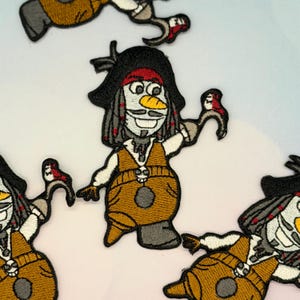 May include: Embroidered patches featuring a cartoon pirate snowman character. The patches have a black pirate hat with a red band, a brown vest, and a white face with a mustache. The snowman is holding a red bird in one hand.