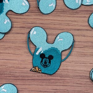 May include: A teal blue embroidered patch of Mickey Mouse's head inside a balloon shaped like Mickey Mouse's ears. The balloon is filled with gold confetti.