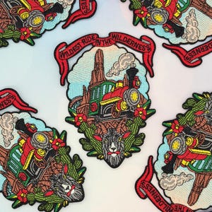 Op de afbeelding: Een geborduurde patch met een rode en groene stoomlocomotief die door een woestijnlandschap met cactussen rijdt, een geitenkop aan de onderkant en een rode banner bovenaan met de tekst Wildest Ride In The Wilderness in zwarte letters.