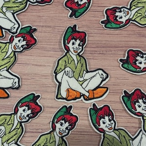 May include: Embroidered patches featuring Peter Pan in a seated pose. The patches depict Peter Pan with red hair, a green hat, and a green outfit. The patches are surrounded by a white border and are scattered across a wooden surface.