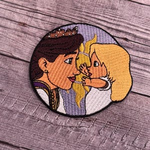May include: Embroidered patch featuring a woman with a crown and a young girl. The woman has brown hair and is wearing a purple and gold dress. The girl has blonde hair and is wearing a white dress. The background is a purple and yellow swirl.