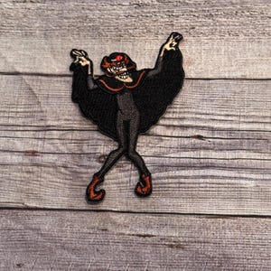 Puede incluir: Un parche bordado con un personaje de dibujos animados con un sombrero rojo y una capa negra, bailando con los brazos en alto.