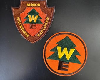 May include: Two embroidered patches featuring a green pine tree with a yellow "W" and "E" inside. The larger patch is brown with the text "Senior Wilderness Explorer". The smaller patch is orange.