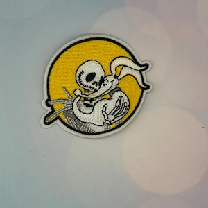 May include: A circular embroidered patch featuring a white skeleton character with a round head and a white ghost dog with long ears. The figures are set against a solid yellow background with a thick black border, creating a high-contrast design.