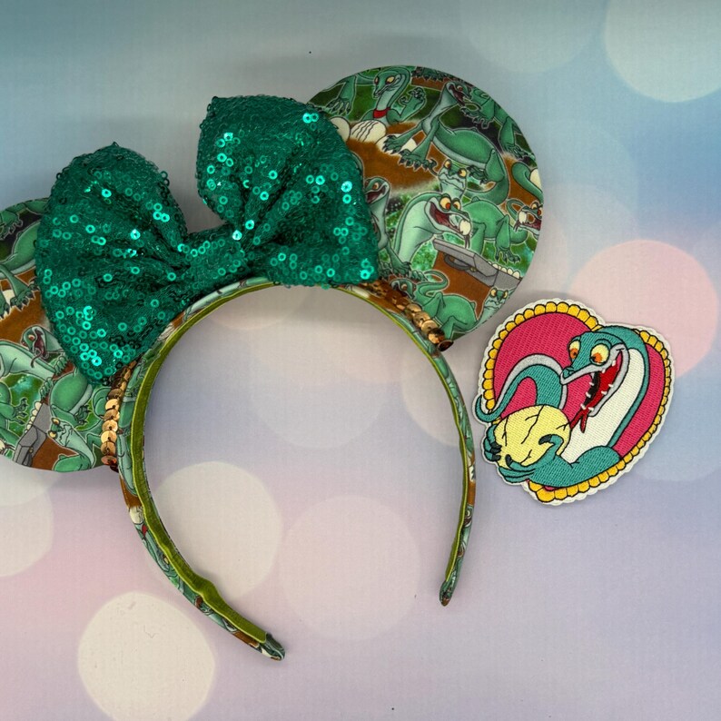May include: Green sequined bow headband with matching ears featuring a cartoon dinosaur print. A heart-shaped patch with a dinosaur design is also included. The headband is a fun accessory for themed events.