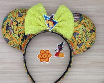 Op de afbeelding: Gele en zwarte muisoren met een gele pailletten strik. De oren hebben een cartoonfiguur met een geel en zwart pak en een rood atoomsymbool. De strik heeft een cartoonfiguur met een geel en zwart pak erop.