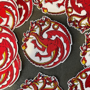 May include: Embroidered patches featuring a red dragon with yellow accents on a white circular background. The dragon has outstretched wings and a flame. The patches are scattered on a dark gray surface.