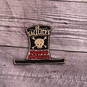 May include: Embroidered patch featuring a black top hat design with the text "DR. FACILIER'S" above a skull and crossbones, and "VOODOO EMPORIUM" in red. The patch has a black border and is set against a wood grain background.