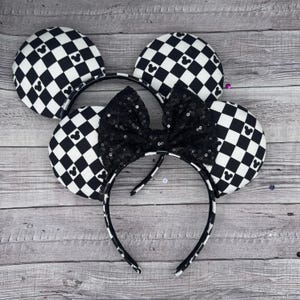 Puede incluir: Orejas de ratón a cuadros en blanco y negro con un lazo de lentejuelas negras. Las orejas están unidas a una diadema negra.