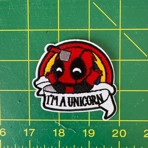 K&ouml;nnte beinhalten: Gestickter Aufn&auml;her mit einer roten und schwarzen Comicfigur mit einem Regenbogen dahinter. Die Figur tr&auml;gt eine wei&szlig;e Maske und der Text "I'm a unicorn" ist auf einem wei&szlig;en Banner geschrieben.