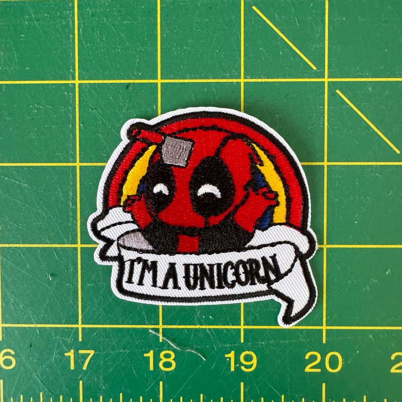 K&ouml;nnte beinhalten: Gestickter Aufn&auml;her mit einer roten und schwarzen Comicfigur mit einem Regenbogen dahinter. Die Figur tr&auml;gt eine wei&szlig;e Maske und der Text "I'm a unicorn" ist auf einem wei&szlig;en Banner geschrieben.