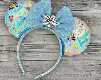 May include: A headband with mouse ears featuring a light blue sequined bow and a "Birthday Princess" emblem. The ears are decorated with a colorful cartoon print of amusement park characters and treats.