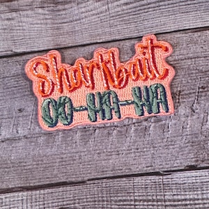 May include: An embroidered patch in coral, featuring the words "Shark Bait" in orange script above "OO-HA-HA" in green block letters. The patch has a pink border and is set against a grey wood-grain background.