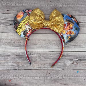 May include: A pair of mouse ears with a gold sequin bow and ear designs featuring scenes from the movie "Bedknobs and Broomsticks". The headband is red with a patterned design, and the ears have a colorful movie poster print.