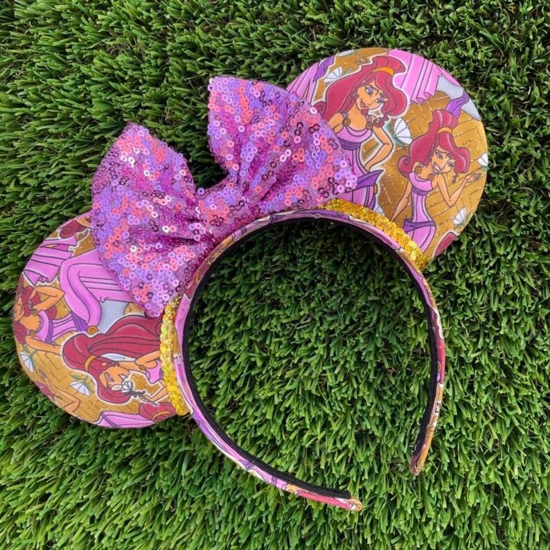 K&ouml;nnte beinhalten: Ein Paar Micky-Maus-Ohren mit einer gro&szlig;en, paillettenbesetzten lila Schleife. Die Ohren haben ein gold-rosa Design mit einer Zeichentrickfigur. Das Stirnband ist lila mit goldenem Besatz.