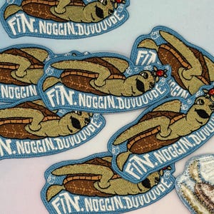 May include: Embroidered patches featuring a cartoon turtle with the text "FIN. NOGGIN. DUUUUUDE" in a light blue bubble design. The turtle has a brown shell and a green body. The patches are likely for clothing or accessories.