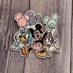 May include: An embroidered patch featuring Mickey Mouse, a blue and white dog, and a brown and white dog. The characters are holding colorful balloons with Mickey Mouse in the center.