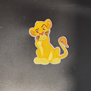 Puede incluir: Un parche bordado de planchado con un cachorro de león de dibujos animados sentado con una expresión seria. El cachorro de león es amarillo con una melena marrón, una nariz rosa y una cola marrón.