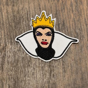 Op de afbeelding: Een geborduurde strijkpatch met de boze koningin uit Sneeuwwitje. De patch is wit met een zwart-witte afbeelding van de boze koningin die een gele kroon draagt. Haar ogen zijn paars en ze heeft een strenge uitdrukking.