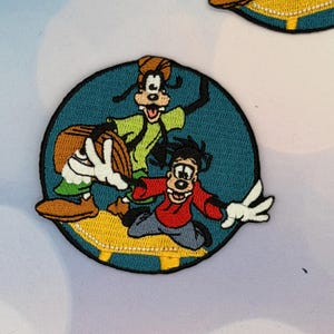 Pode incluir: Patch bordado com Pateta, Max e outro personagem em um círculo verde-azulado. Pateta está vestindo uma camisa verde e um chapéu marrom. Max está de camisa vermelha. O patch tem uma borda amarela.