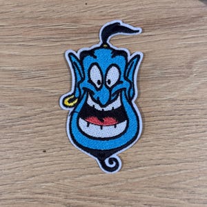May include: A blue Genie patch with a smiling expression, white eyes, and a black beard. The patch has a yellow earring and a black topknot. The patch is outlined in white and is on a wooden surface.