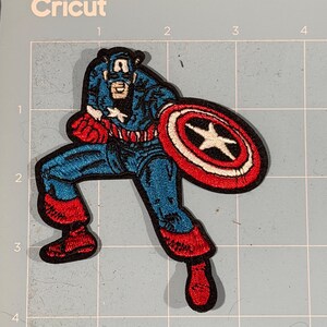 May include: Embroidered Captain America patch in a dynamic pose, holding his shield. The patch features blue, red, white, and black colours. The design is detailed, with visible stitching and a classic comic book style. The patch is likely for clothing or accessories.