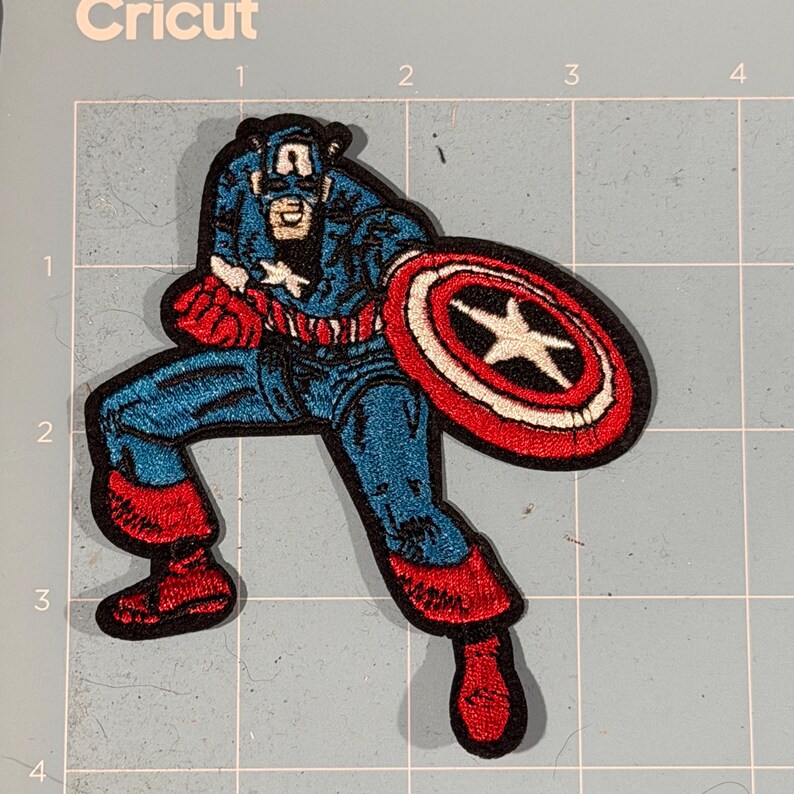 May include: Embroidered Captain America patch in a dynamic pose, holding his shield. The patch features blue, red, white, and black colours. The design is detailed, with visible stitching and a classic comic book style. The patch is likely for clothing or accessories.
