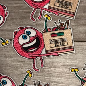 May include: Embroidered patch featuring a pink cartoon character with blue eyes and a wide grin, holding a cardboard box. The box contains flowers and other items. The patch has a white border and is set against a wood grain background.