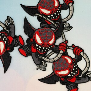 以下が含まれることがあります： 赤と黒の配色で描かれた、漫画の悪魔のキャラクターの刺繍パッチ。悪魔の顔にはクモの巣のような模様があり、不気味な笑みを浮かべている。パッチには黒い翼と腕が付いている。