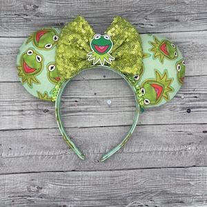 Puede incluir: Orejas de ratón verdes y blancas con un lazo de lentejuelas verdes y una aplicación de fieltro de Kermit la Rana. Las orejas están impresas con un patrón de la cara de Kermit la Rana.