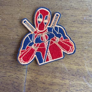 May include: Red and black embroidered patch of Deadpool with his hands in a heart shape. The patch has a gold outline.