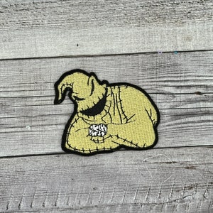 May include: A yellow embroidered patch of the character Oogie Boogie from the movie The Nightmare Before Christmas. He is holding a bag of hearts in his hands.