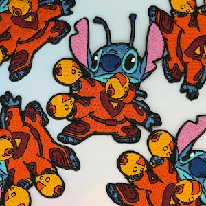 May include: Embroidered patches featuring a blue alien character with large ears and eyes, wearing an orange spacesuit. The patches have a detailed design with various colors, including orange, yellow, blue, and pink. The patches are likely for clothing or accessories.