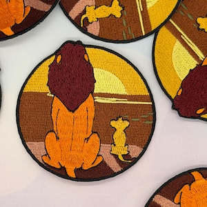 Puede incluir: Parches bordados circulares que representan un león y su cachorro al atardecer. El león es naranja con una melena granate, y el cachorro es amarillo. El fondo incluye un sol amarillo y un paisaje marrón.