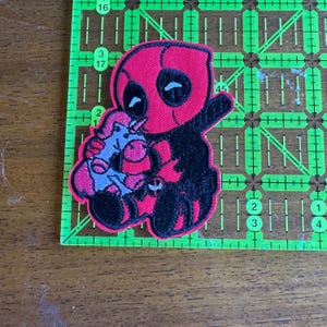 Puede incluir: Un parche bordado rojo y negro de Deadpool sosteniendo un unicornio rosa.