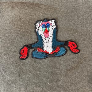 May include: Embroidered patch featuring a cartoon baboon in a seated yoga pose. The baboon has a blue body, white fur, red hands and feet, and a red and green face. The patch is outlined in black.