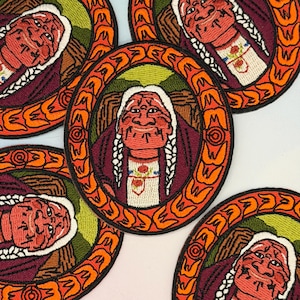 May include: Embroidered patches depicting a person with white braids, wearing a burgundy top and a white shirt with floral embroidery. The portrait is set within a circular frame with orange and brown decorative elements. The patches are arranged on a white surface.