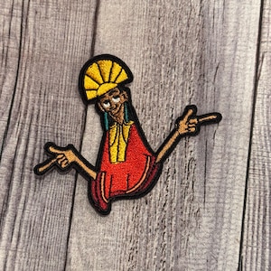 May include: Embroidered patch depicting a cartoon character in a red and yellow outfit, with arms outstretched and fingers pointing. The character has a yellow headdress and is outlined in black. The patch is on a grey wooden surface.