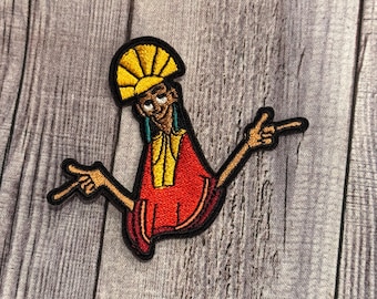May include: Embroidered patch featuring a cartoon character in a red and yellow outfit, with arms outstretched and fingers pointing. The character has a yellow headdress and is outlined in black. The patch is on a gray wooden surface.