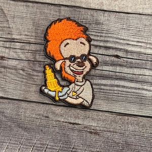 May include: An embroidered patch featuring a cartoon character with orange hair, wearing glasses and a white shirt. The character is holding a yellow object that resembles a corn cob.