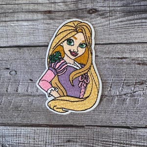 Op de afbeelding: Geborduurde patch met een cartoonprinses met lang blond haar, groene ogen en een kikker op haar schouder. Ze draagt een paarse jurk met roze mouwen en een roze strik. De patch heeft een witte rand.
