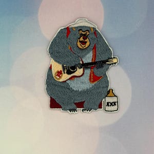 May include: An embroidered patch featuring a grey bear wearing a white hat and a red vest, playing a light-coloured acoustic guitar. Beside the bear sits a white jug with the letters XXX printed in black on the front of the container.