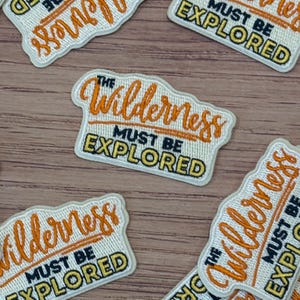 May include: Embroidered patches with the text "The Wilderness Must Be Explored" in orange and black. The patches have a rectangular shape with a white border and are scattered on a wooden surface.