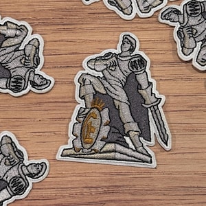 May include: Embroidered patch featuring a knight in silver armor, holding a sword and shield with a gold emblem. The knight is posed on a stone structure. Several patches are scattered on a wooden surface.