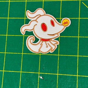 Könnte beinhalten: Ein weißer gestickter Patch von Null, dem Geisterhund aus Nightmare Before Christmas, mit roten Augen und einem gelben Halsband. Der Patch befindet sich auf einer grünen Schneidematte.