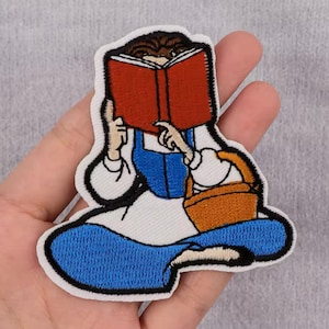 May include: Embroidered iron-on patch featuring a cartoon character sitting with a book in their lap and a basket in their hand. The character is wearing a white shirt, blue overalls, and blue pants. The book is red and the basket is orange.